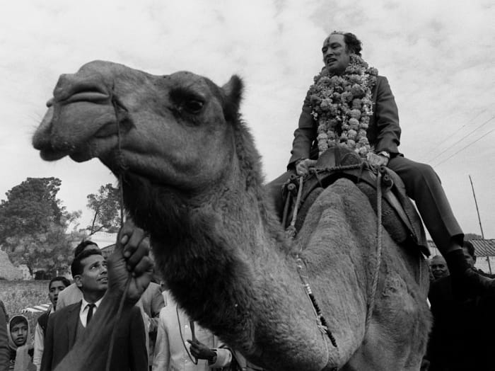 Pierre Trudeau, with a garland around his neck and a Hindu greeting symbol in paste on his forhead, rides a camel in the village of Benares, India, where he dedicated a water well (Jan 12, 1971).