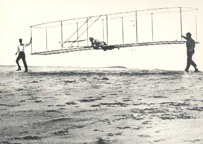 Historic photo of the Wright brothers' third test glider being launched at Kill Devil Hills, North Carolina, on October 10, 1902. Wilbur Wright is at the controls, Orville Wright is at left, and Dan Tate (a local resident and friend of the Wright bro