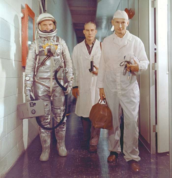 John Glenn, suited-up pre-flight, is seen carrying the portable ventilation unit. Photo courtesy of NASA.