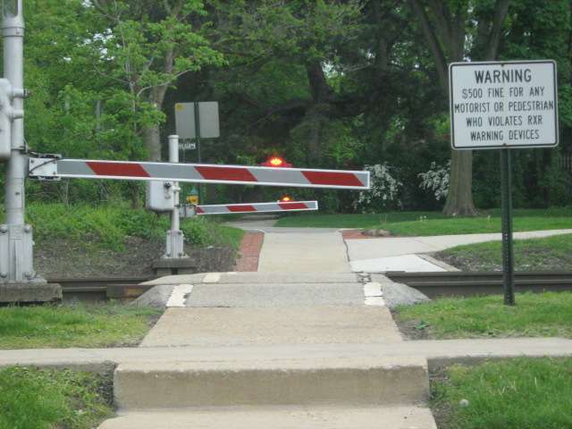 A pedestrian crosswalk near Downtown Hinsdale with the gate down, lights flashing, as the Metra train nears from Chicago