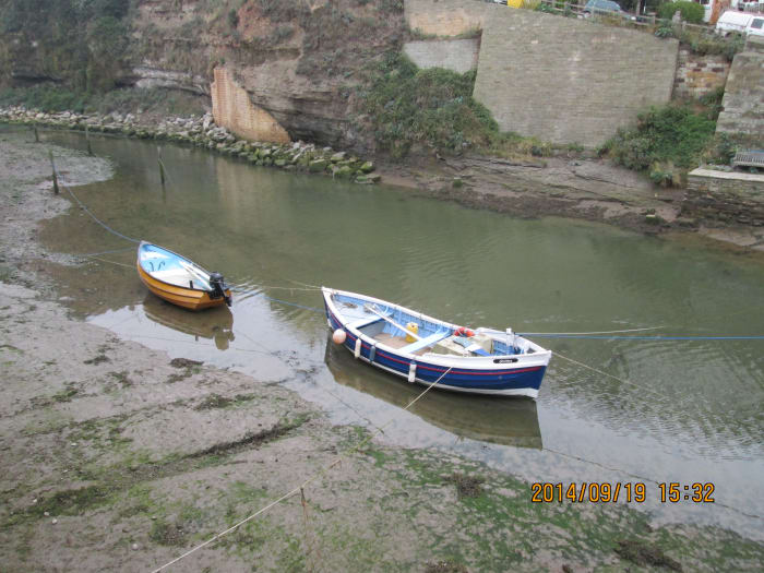 A Yorkshire coble and smaller craft at high tide on the creek, seen from the riverside walk near the small, narrow wooden footbridge