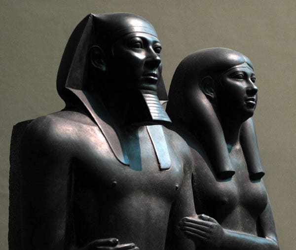 The two figures stand side-by-side, gazing into eternity. He represents the epitome of kingship and the ideal human male form. She is the ideal female. He wears the Nemes on his head, a long artificial beard, and a wraparound kilt with central tab, a