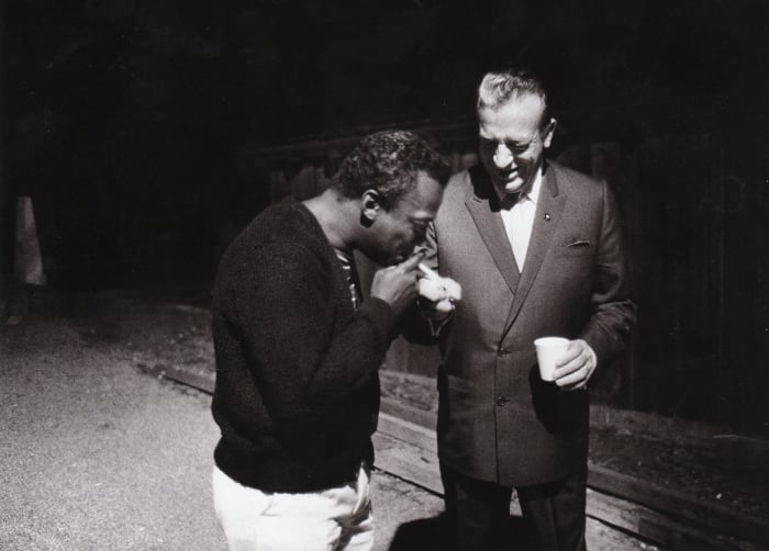 Miles Davis having a cigarette lit by Harry James on the backstage at the Monterey Jazz Festival 1963.