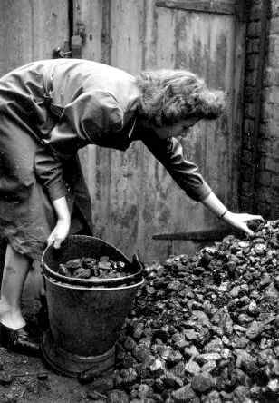 A home help fills a coal bucket with coal during a visit to the house of an invalid, so that there will be a plentiful supply of fuel within easy reach.