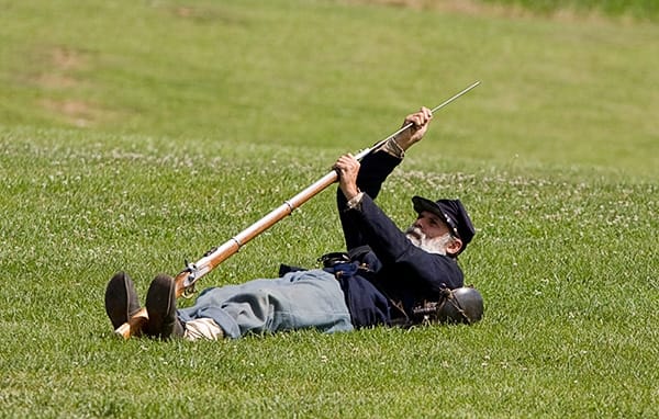 A Living Historian loads his musket from the Prone Stance. He is on his back, balances the firearm between his feet, and is about to ram the cartridge down the barrel