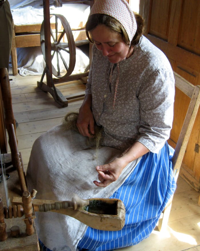 Stripes seem to be popular for the gathered, long skirts. This woman wear a patterned cap that ties under her chin, instead of the plain white head covering. Her apron is a rough weave, so possibly flax or linen. She is spinning fiber.