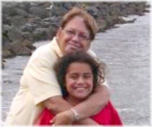 and a young girl who appears to be her granddaughter. The woman has short, reddish, wavy hair and a friendly smile. She is wearing a white blouse. She is hugging her smiling granddaughter who has long, dark curly hair and is wearing a red T-shirt.