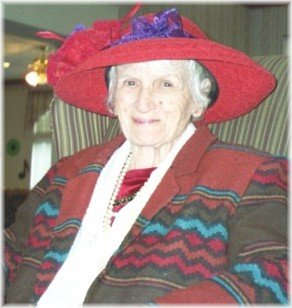 She is slim and petite with a pleasant smile and mischievous eyes. She has snowy white hair and is wearing a big red hat and a colorful jacket. She seems to be a lady with a lot of character.