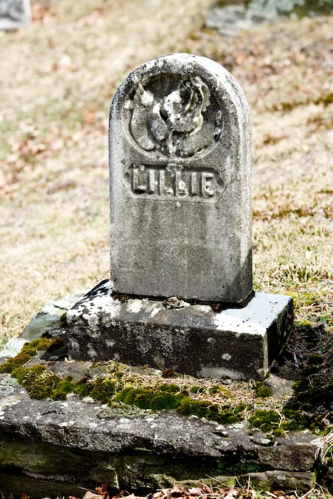 This old domed tablet headstone sets on a base and simply has the name "Lillie". The rosebud carving at the top indicates a young life. The small size of the stone further suggests a very young child or baby.