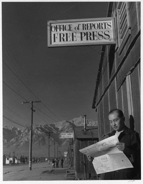 Roy Takeno reading paper in front of office, by Ansel Adams
