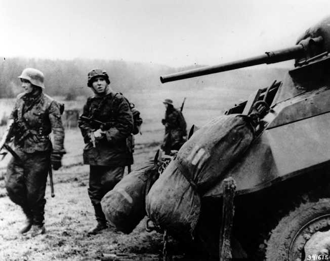 German troops passing by an abandoned American armored car as American troops quickly retreat toward safer ground.