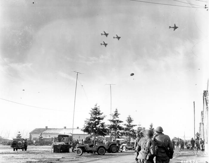 Photo taken by a U.S. Army Signal Corps photographer on December 26, 1944, in Bastogne, Belgium as troops of the 101st Airborne Division watch C-47s drop supplies.
