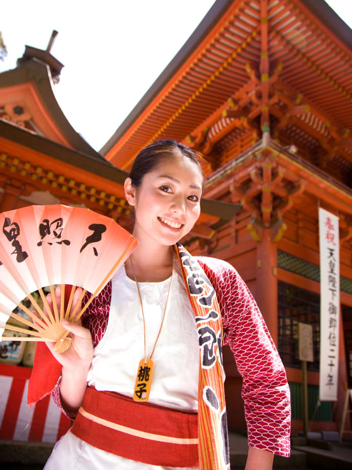 Momoko Tani standing outside of a temple perhaps? She is holding one of those small fans that has the ability to cool you off.