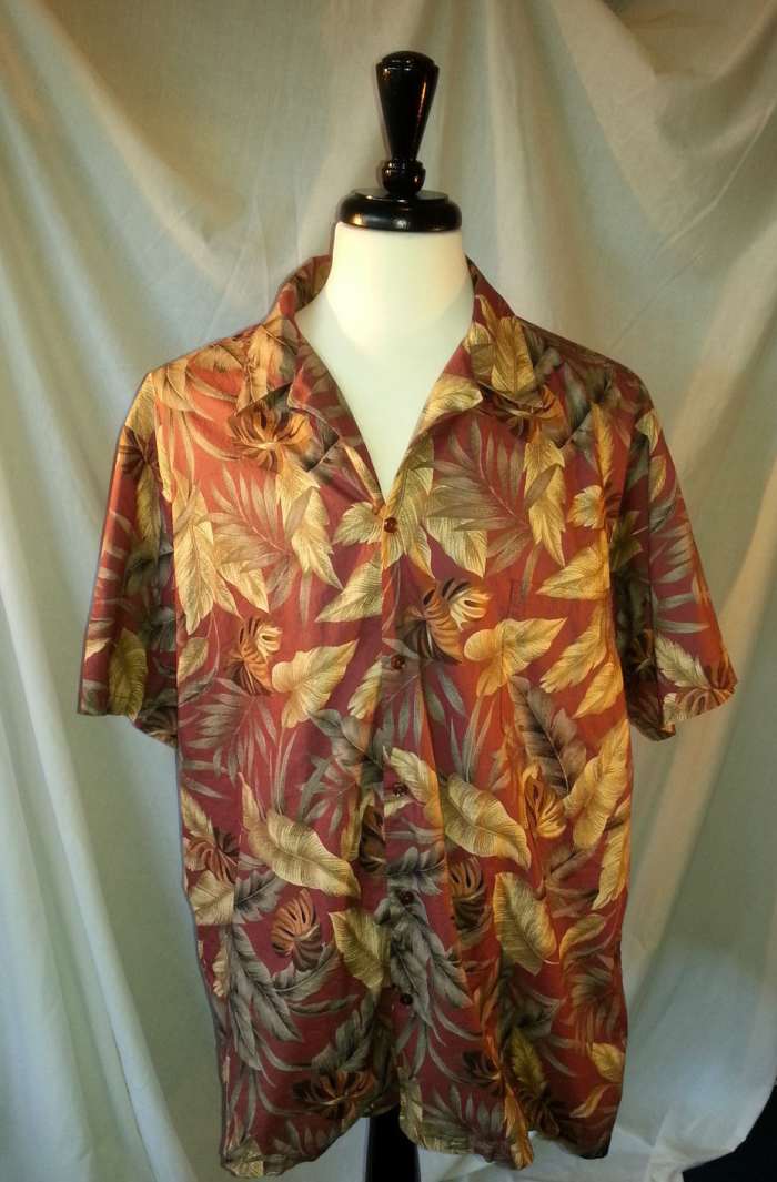 This Hilo Hattie shirt features a casual yet fun sophisticated Hawaiian print with a brick red background with a variety of gold and green leaves.