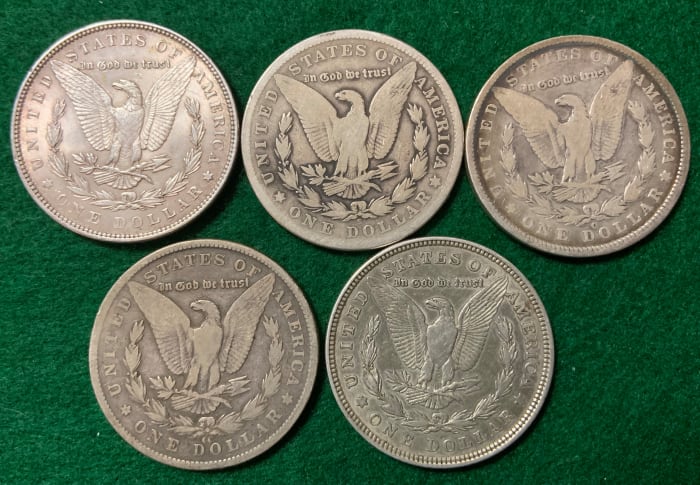 Five mint marks of the Morgan dollar below eagle. Upper-left: no mint mark for Philadelphia. Middle-top: S for San Francisco. Top-right: O for New Orleans. Bottom-left: CC for Carson City. Bottom-right: D for Denver (note how small it is).