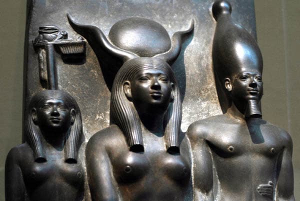 Men-Kau-Re stands on Hathor's left with his left foot advanced. His remarkably individualized facial feature - the prominent eyes, fleshy nose, moustache and protruding lower lip are all characteristics recognizable in other presentations of the King