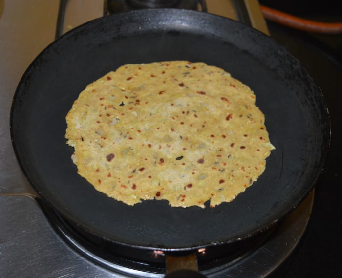 Add a few drops of oil or ghee on the top side. Spread it evenly. Let the other side of the paratha become golden brown. Remove it and place it on a plate. Similarly, cook all the raw parathas and stack them one over the other.