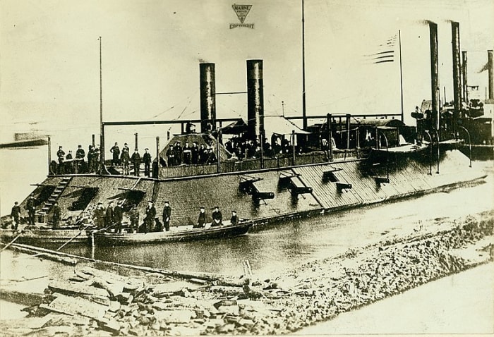 Photograph of an USS Cairo ironclad gunboat with sailors onboard. Two smaller rowboats are in the foreground with more sailors in them. In the background are other riverboats whose smoke stacks are visible.
