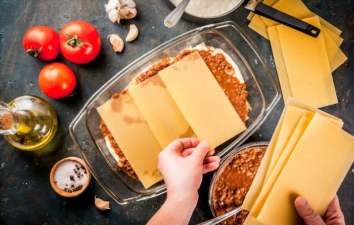 Woman cooking homemade classic lasagna bolognese, on dark blue table; with ingredients, top view copy space, hands in picture