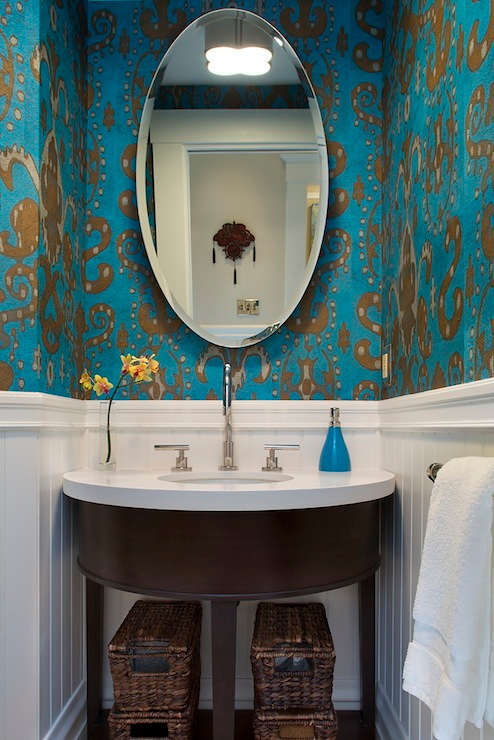 A blue powder room with a half-wall of white beadboard and ikat wallpaper in shiny gold and peacock blue. A demi-lune vanity with white counter sink with sleek faucet sits under a oval mirror in the bathroom.