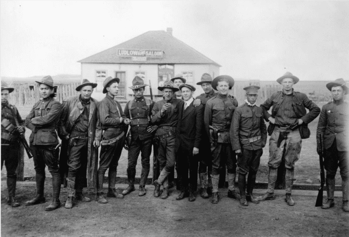 Members of the Colorado National Guard, called in to suppress the UMWA strike against CF&I, pose outdoors with a civilian near the Ludlow Home Saloon in Ludlow, Las Animas County, Colorado. They wear ammunition belts with holsters.