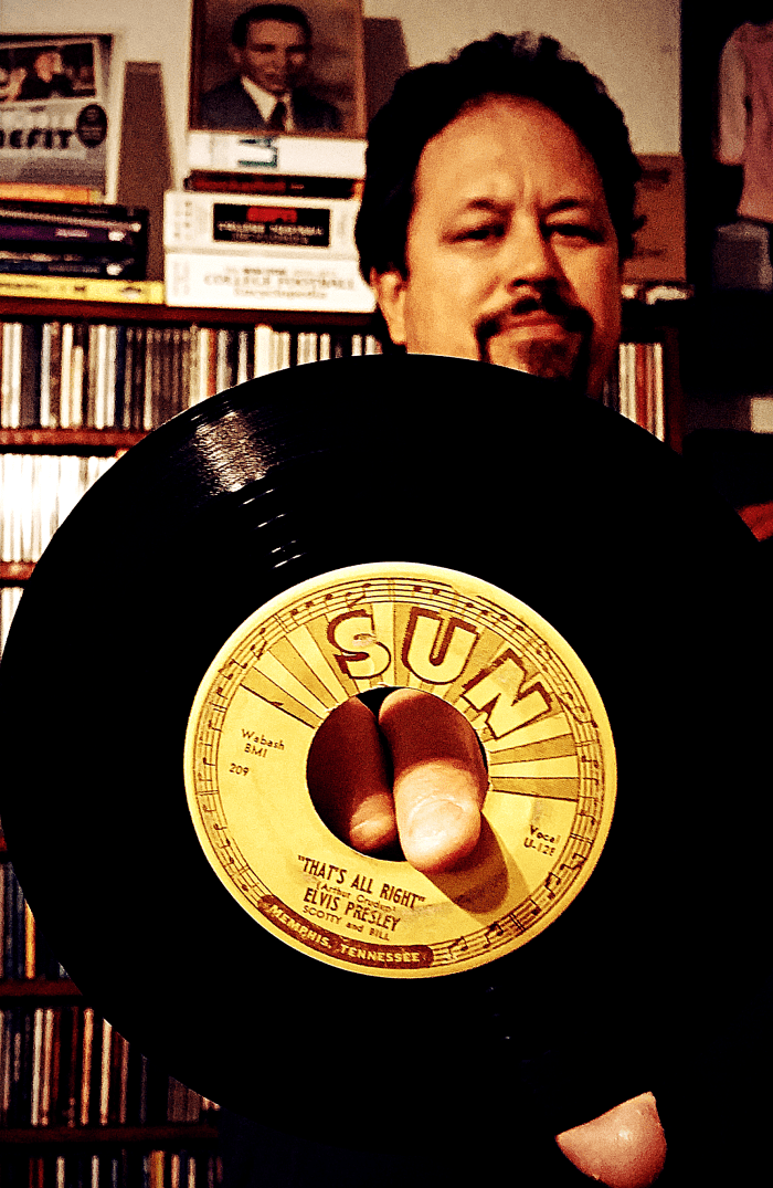 Me holding Elvis' debut single in October 2015, bequeathed to me by my father, a lifelong Presley fan and the young man in the frame behind my head (circa 1951).