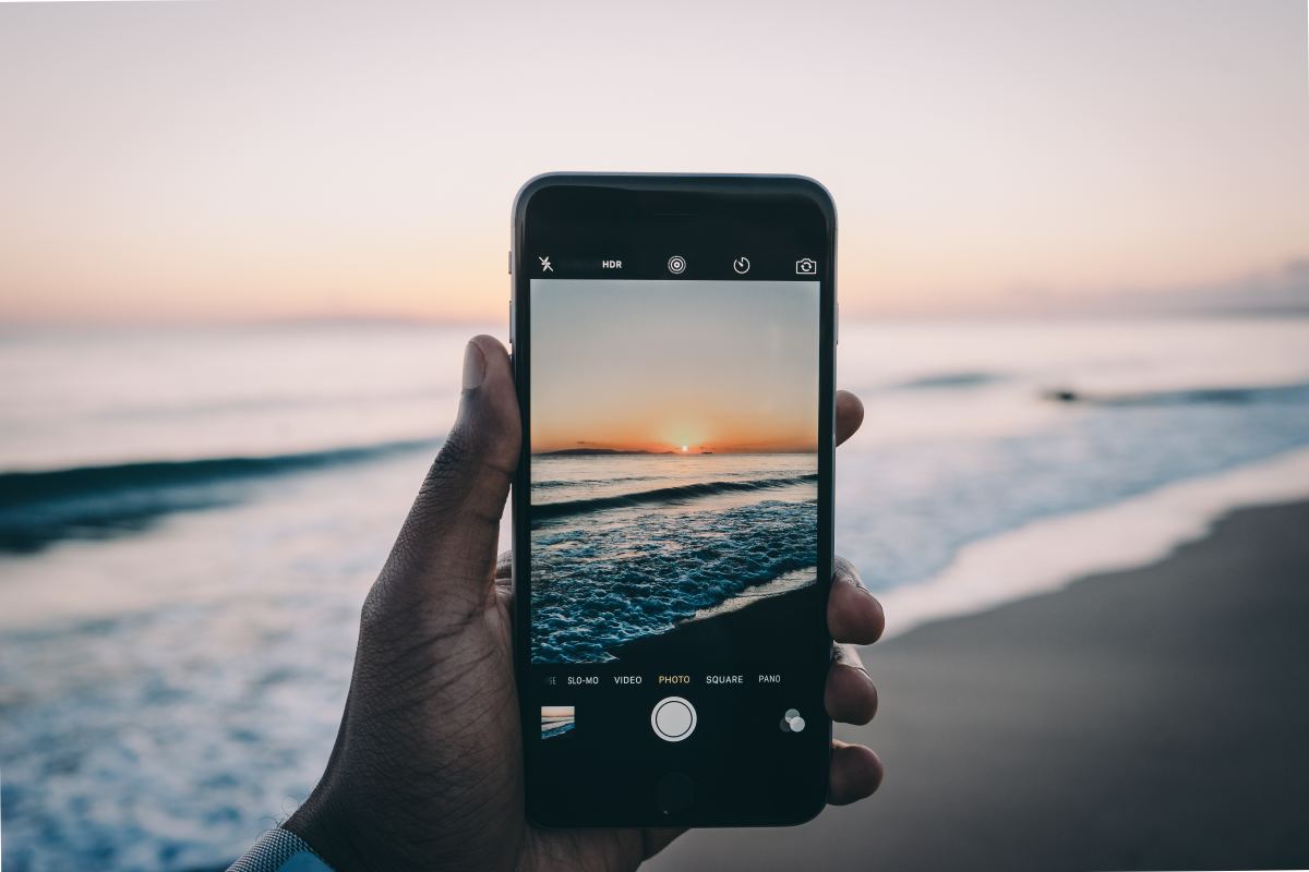  A hand holding a smartphone in the Camera app to take a photo of the beach at sunset.