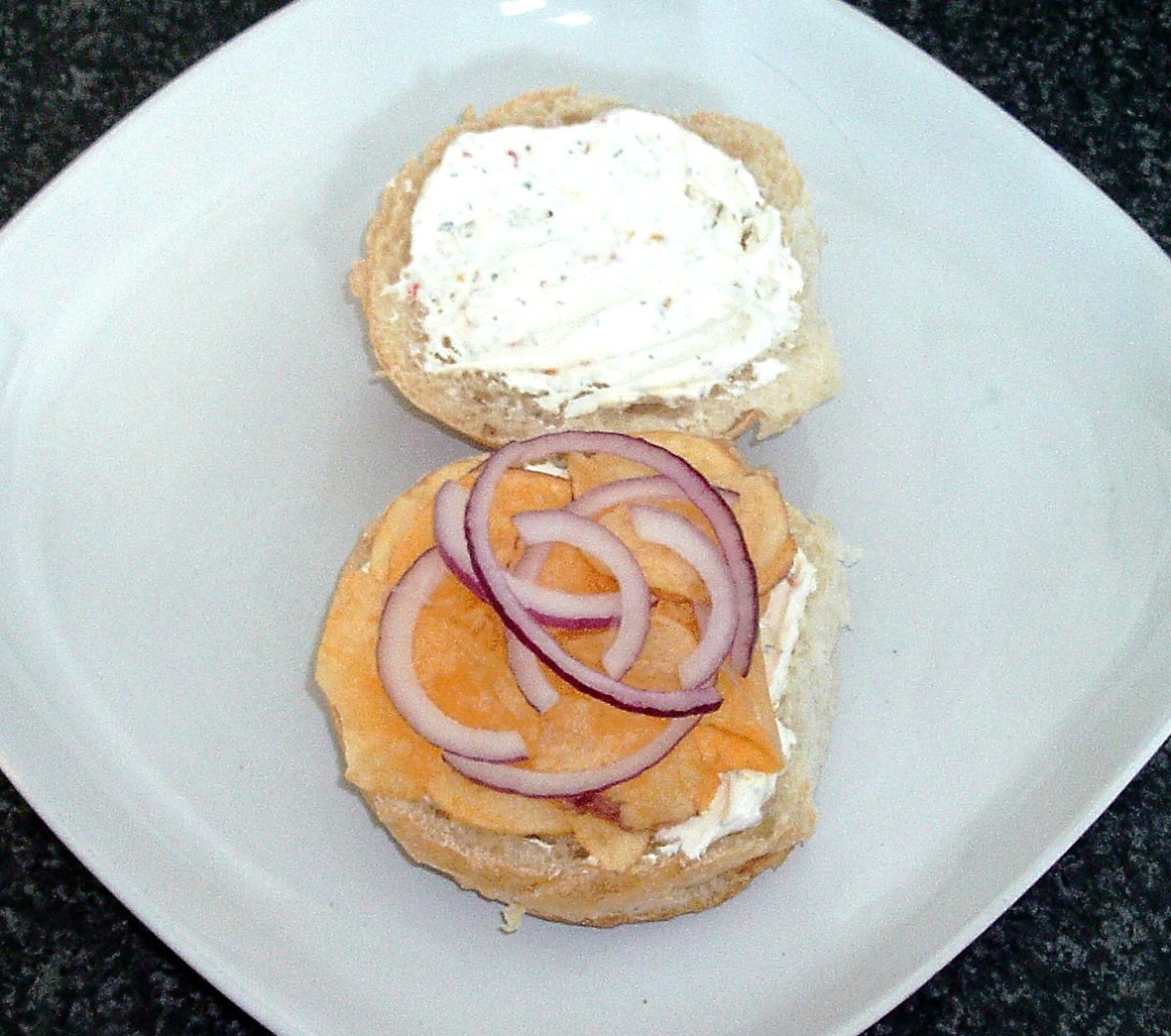 Herb and spice cream cheese is spread on bread roll before cheese and onion crisps and red onion slices are added