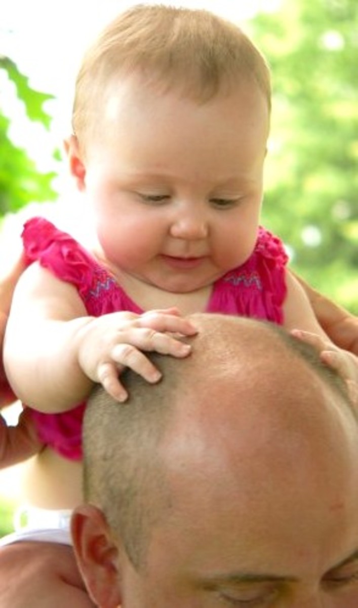 This looks like a little girl wearing a pink top. She has plump round cheeks, and chubby little hands. She seems to be very interested in her Daddy's bald head!