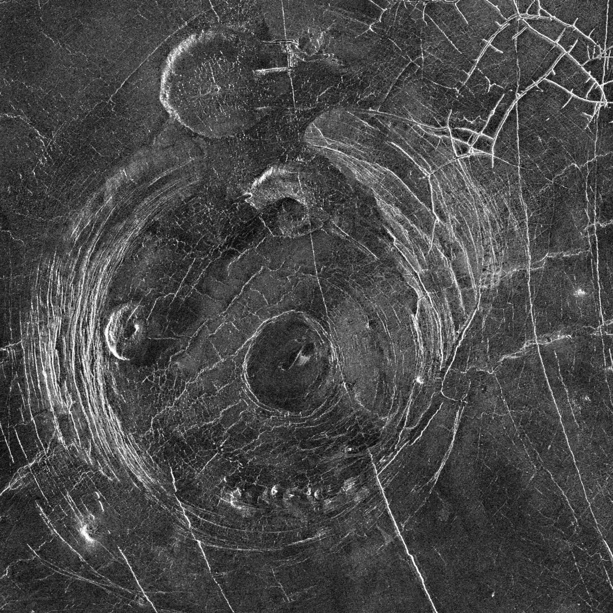 Aine Corona is 200 km (120 miles) in diameter. Above and to the left of the corona are two smaller, rounded 'pancake domes', and top right are fractures through which lava has flowed