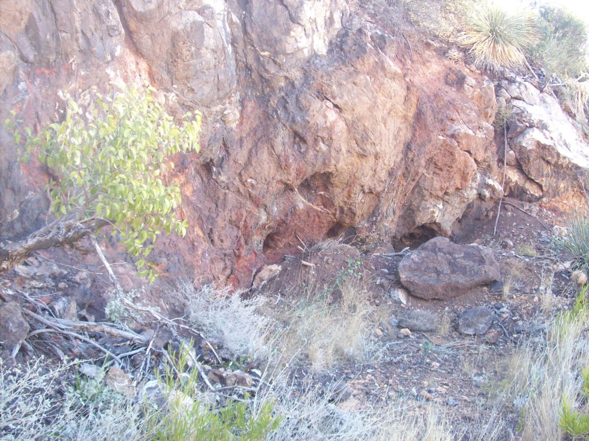 一个巨大的岩石表面，红色的色调，石英条纹和圆形的孔——岩石的硬度在整个表面上是一致的——这些看起来像凿出来的孔。前面是一片空地。
