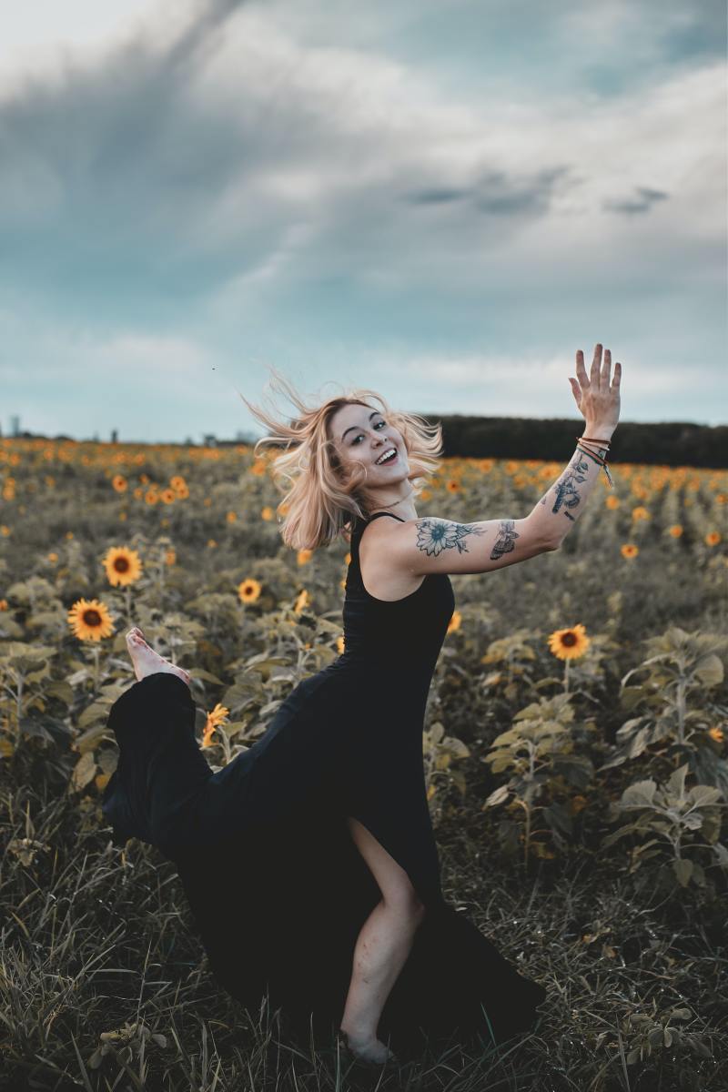 A woman standing in a field of sunflowers while sporting sunflower tattoos on her arm.
