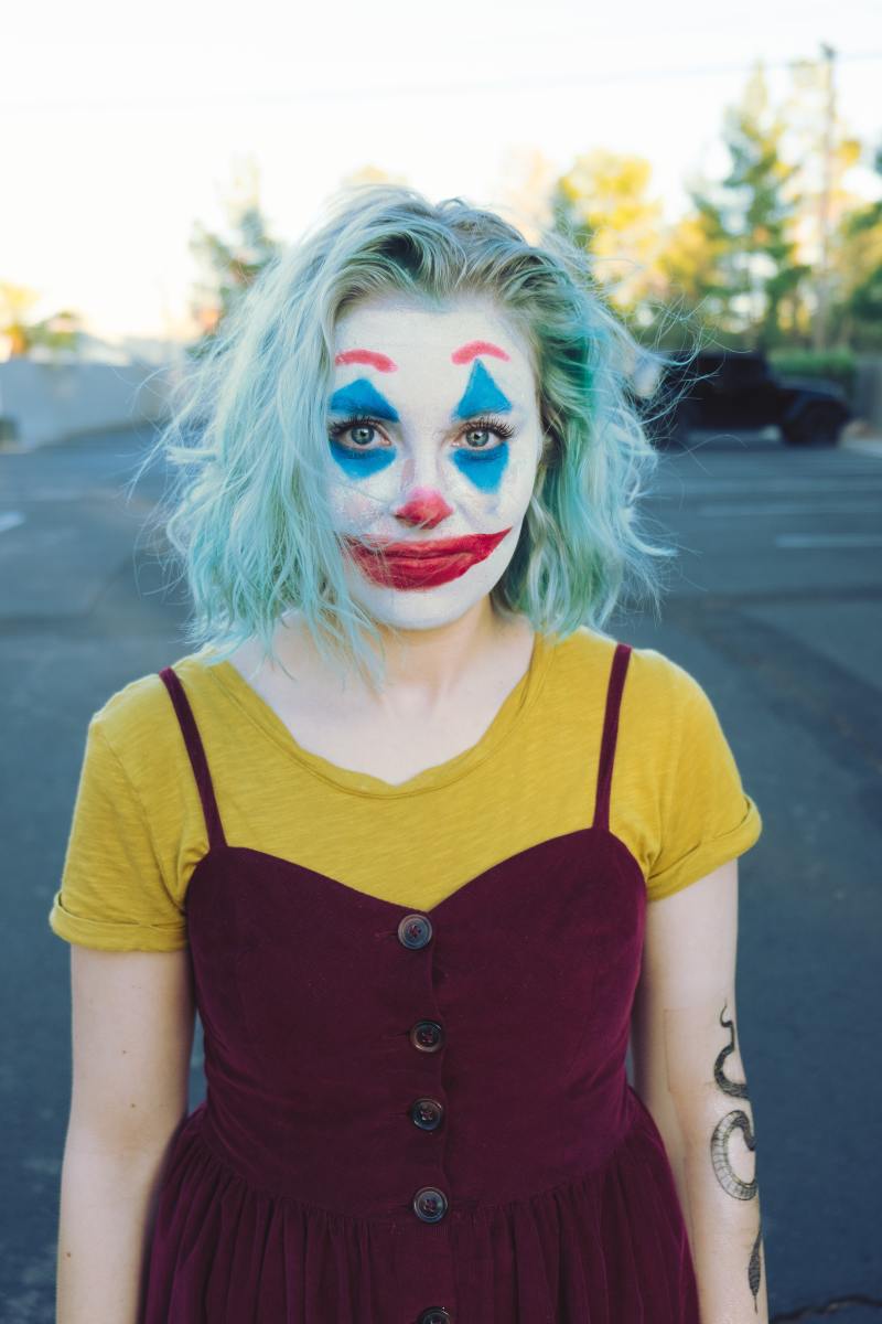 A young lady wearing a clown's face made with facial makeup and a blue-green wig. 