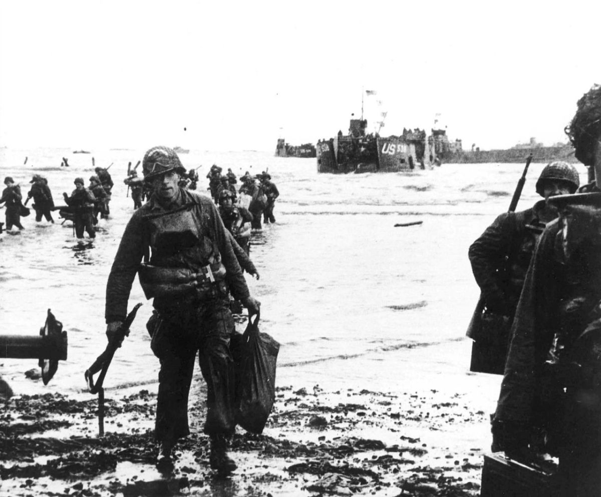 Carrying their equipment, U.S. assault troops move onto Utah Beach almost unopposed. Landing craft can be seen in the background.