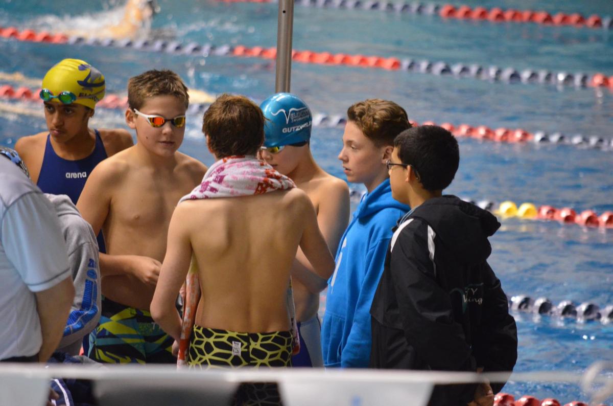 Young swimmers chat between races. Note: swim caps, goggles, and clothes to stay warm between their time racing.
