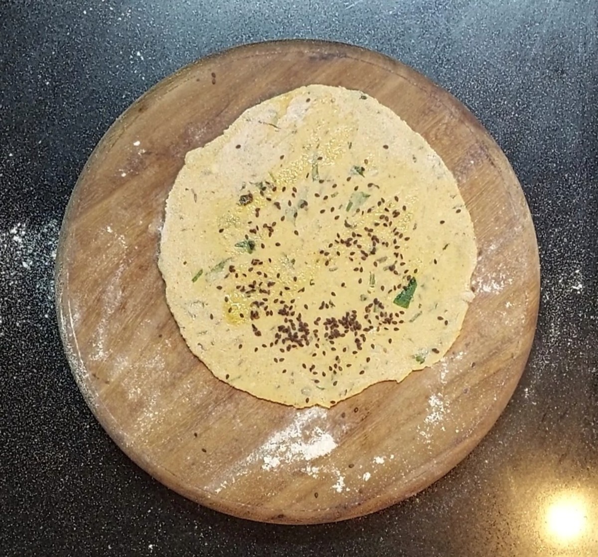 Use a rolling pin to roll the dough into a round paratha. Drizzle a few drops of ghee and spread it over the top. Sprinkle a few sesame seeds.