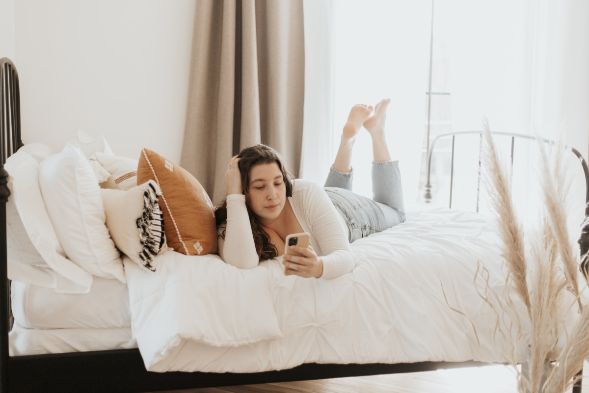  A young woman lying on a bed and using a smartphone to read an article about tips for staying safe while online dating.