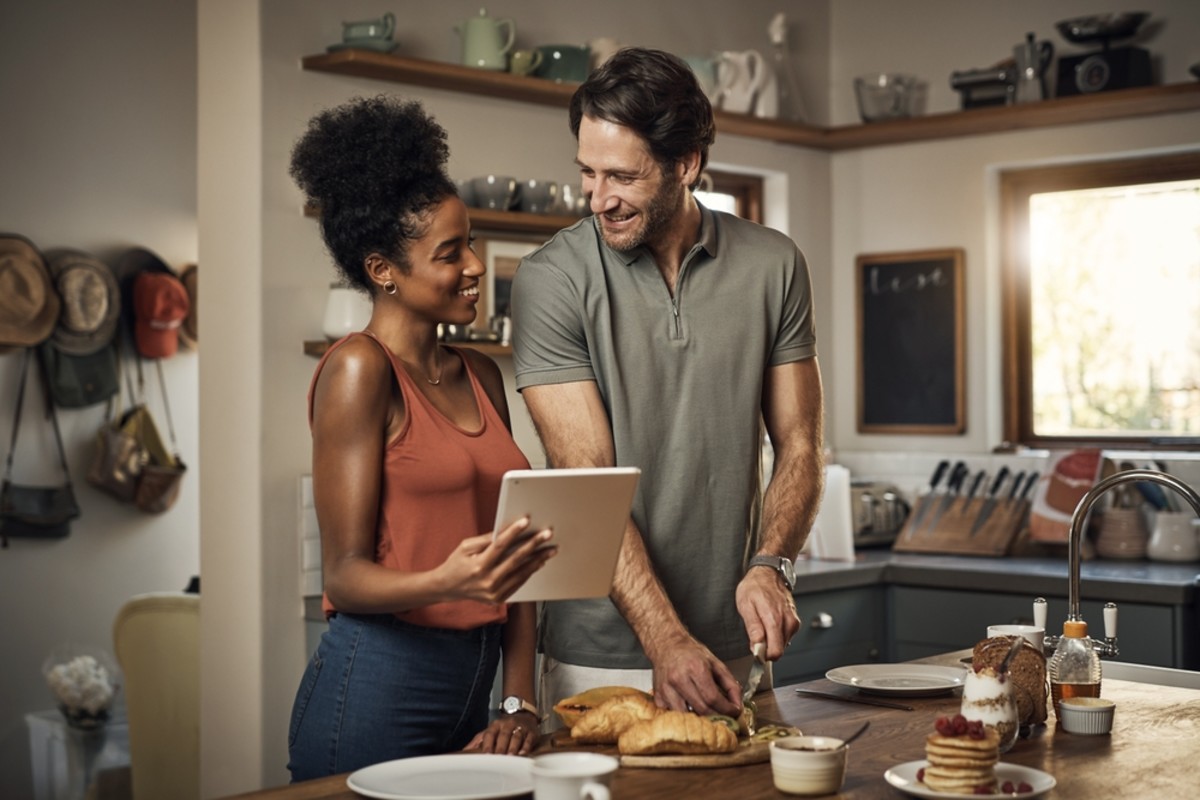  A husband and wife are in the kitchen. The husband is cutting fruit while his wife looks at a tablet with a smile on her face.